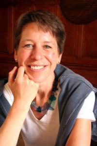 Woman smiling at camera. She wears a white shirt and has a grey jumper draped over her shoulders. She is resting her face on one hand and facing the camera.